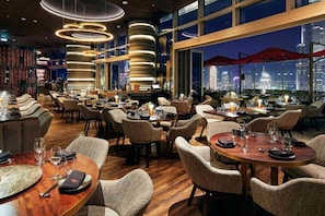 A restaurant with multiple dining tables, chairs, and a city view through large windows.