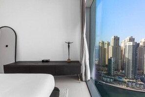 A modern hotel room with a large window, a bed, a bedside table with a lamp, and a view of a cityscape.