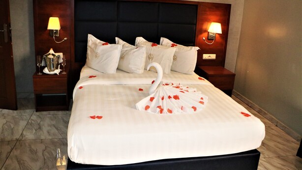 A neatly made bed with a swan design on the bedspread, flanked by two bedside tables with lamps, and a dark headboard. - Jouvence International Hôtel (Yaoundé)