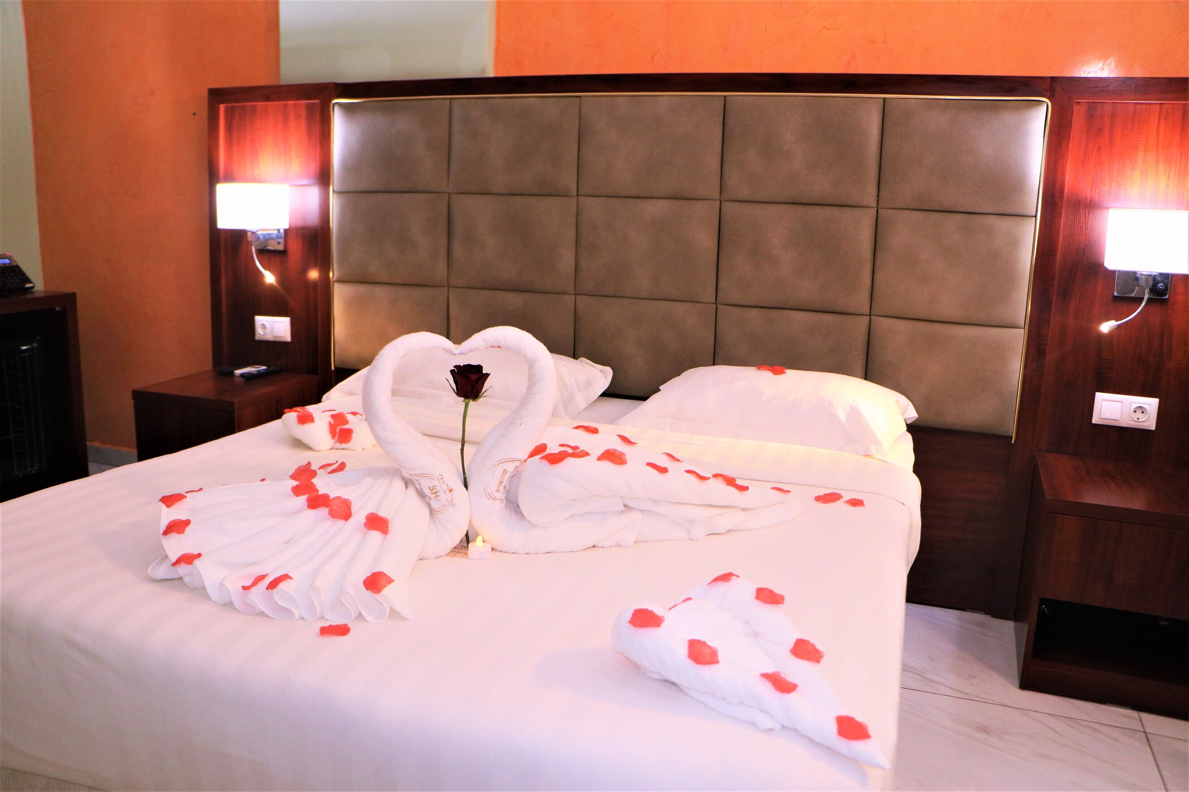A neatly made bed with rose petals and a heart-shaped towel arrangement, a bedside lamp, and a wooden headboard.