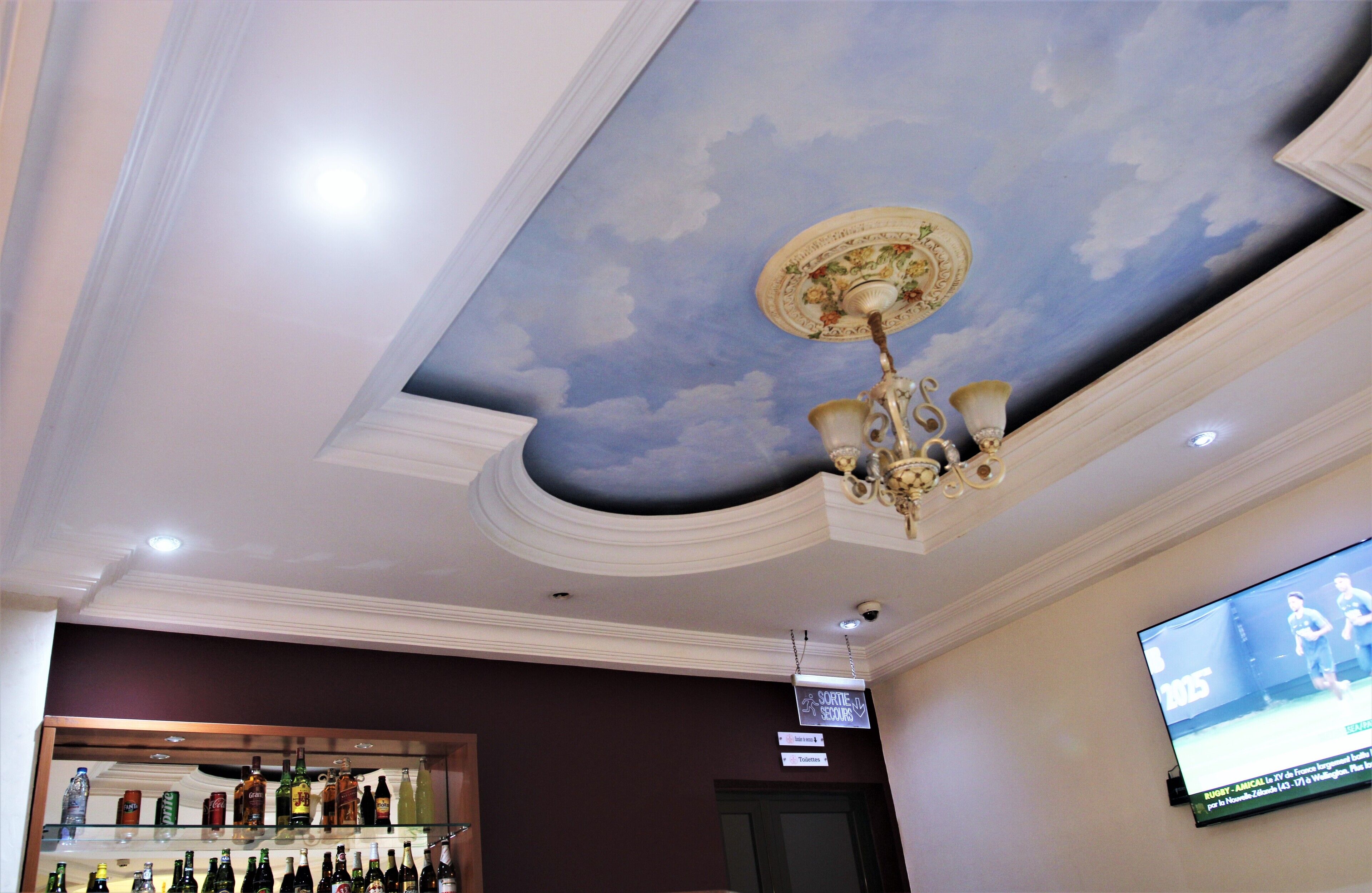 Ein Zimmer mit Himmel- und Wolken- bemaltem Deckenbild, einem Kronleuchter, einem an der Wand montierten Fernseher und einer Bar mit verschiedenen Flaschen.