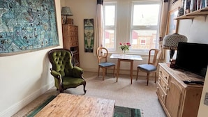 A cozy living room with a green armchair, a wooden coffee table, a dining table with two chairs, a TV on a wooden cabinet, and a large window with curtains. - Apartmentsleeps2-seaside Escape-wifi-parking (Hastings)