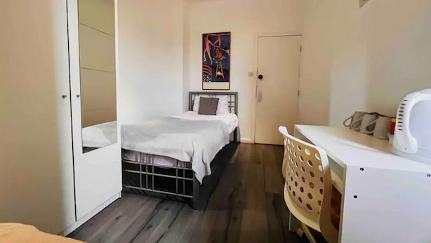 Ein kleiner, kompakter Raum mit Bett, Schreibtisch und Stuhl. Eine Tür befindet sich an einer Seite, ein Fenster an der gegenüberliegenden Wand.