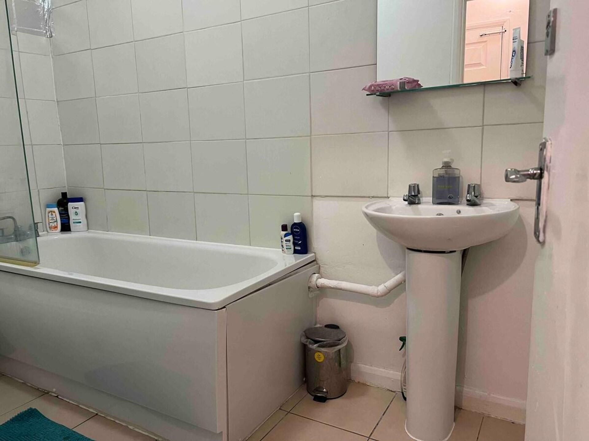 Un baño con bañera, lavamanos y espejo. Hay artículos de aseo en el lavamanos y un cubo de basura al lado.