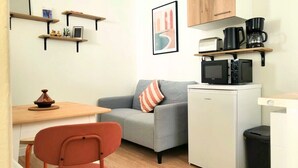 A compact living space with a grey sofa, a small table, a microwave, a coffee maker, and a trash bin.