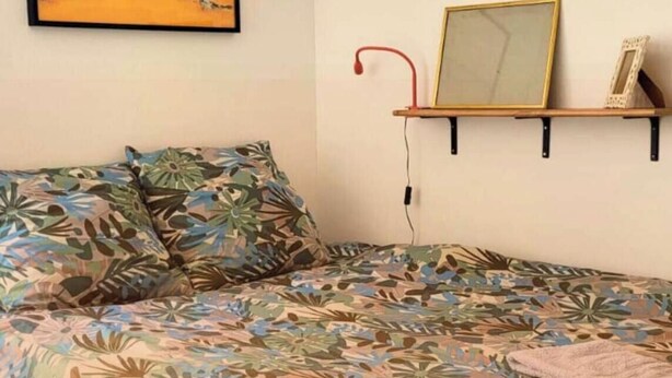 A bed with floral bedding, a bedside lamp, a framed picture, and a shelf with items.