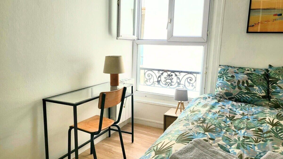A bedroom with a bed, a chair, a small table with a lamp, and a window with a balcony.