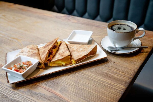 En måltid bestående av en hopvikt quesadilla, en liten skål med salsa och en kopp kaffe, serverat på ett träbord.
