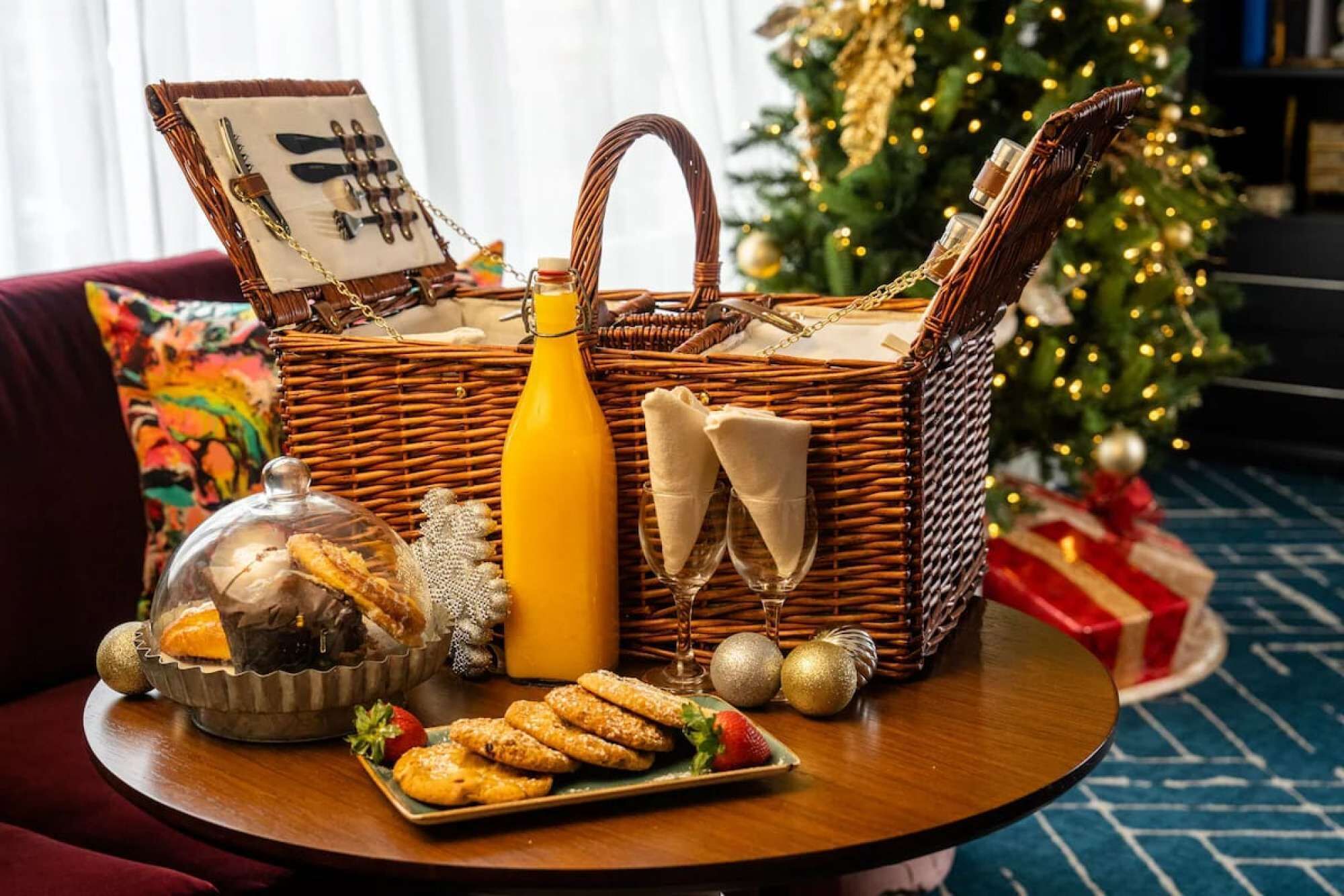 A wicker basket with a yellow bottle, a glass of orange juice, and a plate of pastries on a table. A Christmas tree is in the background.
