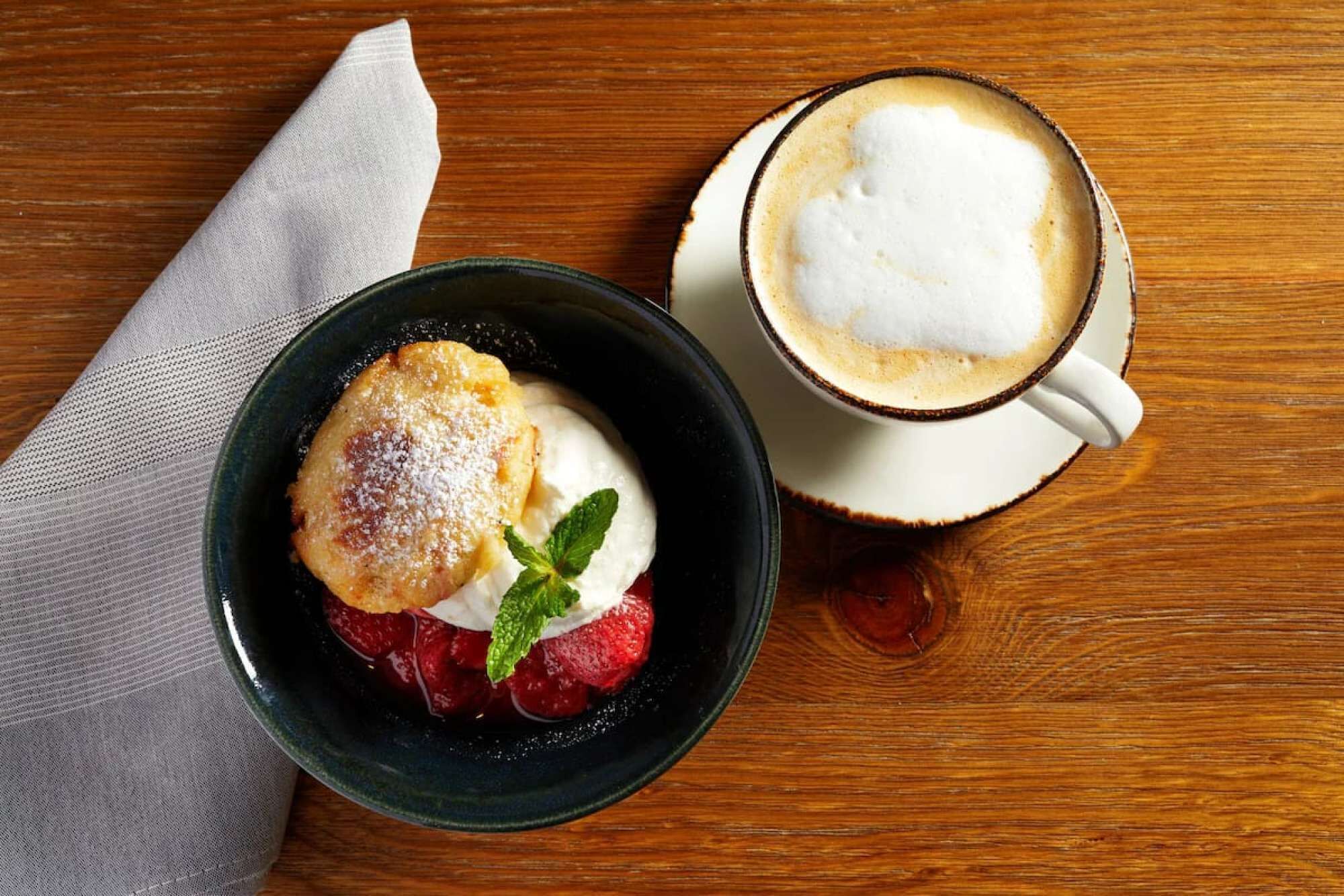 A dessert with a scoop of ice cream, a piece of cake, and a mint leaf on a dark bowl, next to a cup of coffee on a wooden table.