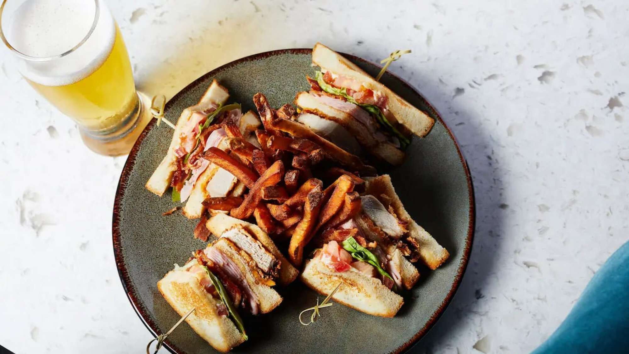 Un plato de sándwiches con tocino y papas fritas, acompañado de una cerveza en una superficie de mármol.