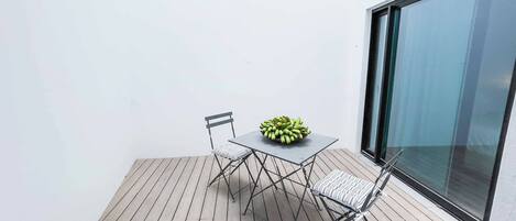 A small outdoor patio with a table and two chairs, a bowl of bananas, and a large window.