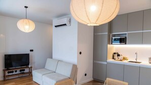 Una sala de estar moderna con un sofá, una mesa de vidrio y un televisor. Cuenta con una zona de cocina con microondas y fregadero.
