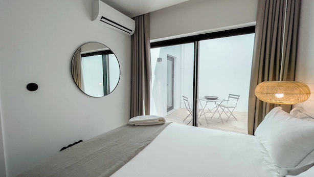 Habitación de hotel con cama, espejo redondo, mesa con sillas y balcón con cortinas.