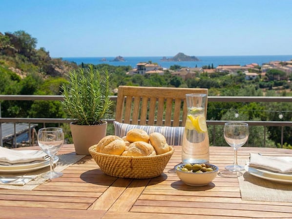 A wooden table set with bread, olives, a glass of water, and a bottle of lemon water, overlooking a scenic coastal view.