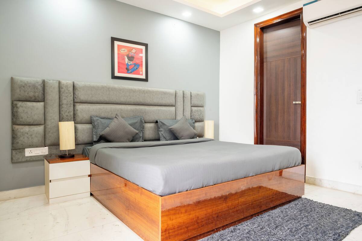 A modern bedroom with a grey upholstered headboard, a wooden bed frame, a grey mattress, a bedside table with a lamp, and a framed picture on the wall.
