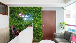 A reception area with a green wall, a reception desk, a grey sofa, and a round table. - Housr 3 BHK Apartment Sector 45 Gurgaon (Gurugram)