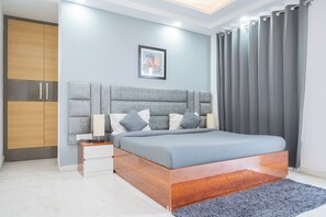 A modern bedroom with a grey upholstered headboard, a wooden bed frame, a bedside table with a lamp, and a grey curtain.