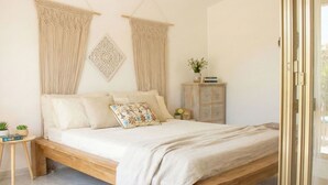 A bedroom with a wooden bed, beige bedding, a small bedside table with a plant, a cabinet, and macramé wall hangings.
