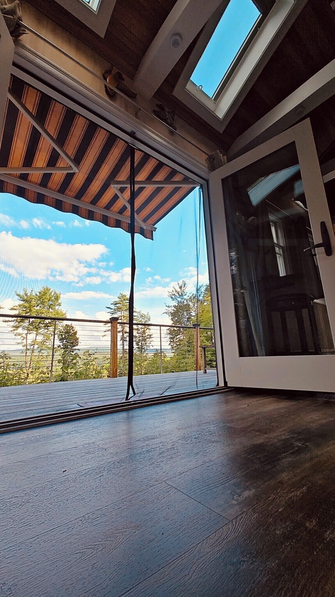 Ein Zimmer mit einer großen Glasschiebetür, die auf eine Außenterrasse führt, einem Oberlicht über der Tür und Blick auf Bäume und Himmel.