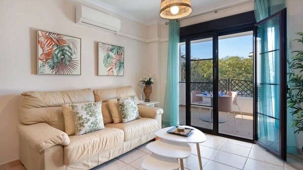 Ein Wohnzimmer mit einem beigefarbenen Sofa, zwei an der Wand befestigten Kunstwerken, einer hängenden Leuchte, einem kleinen weißen Tisch und einer Schiebetür zu einer Außenpatio.