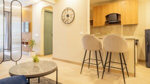 A modern living room with a round coffee table, two bar stools, a clock on the wall, and a kitchen area with wooden cabinets and a black range hood. - Housr 1 BHK Apartment Sector 42 Gurgaon (Gurugram)