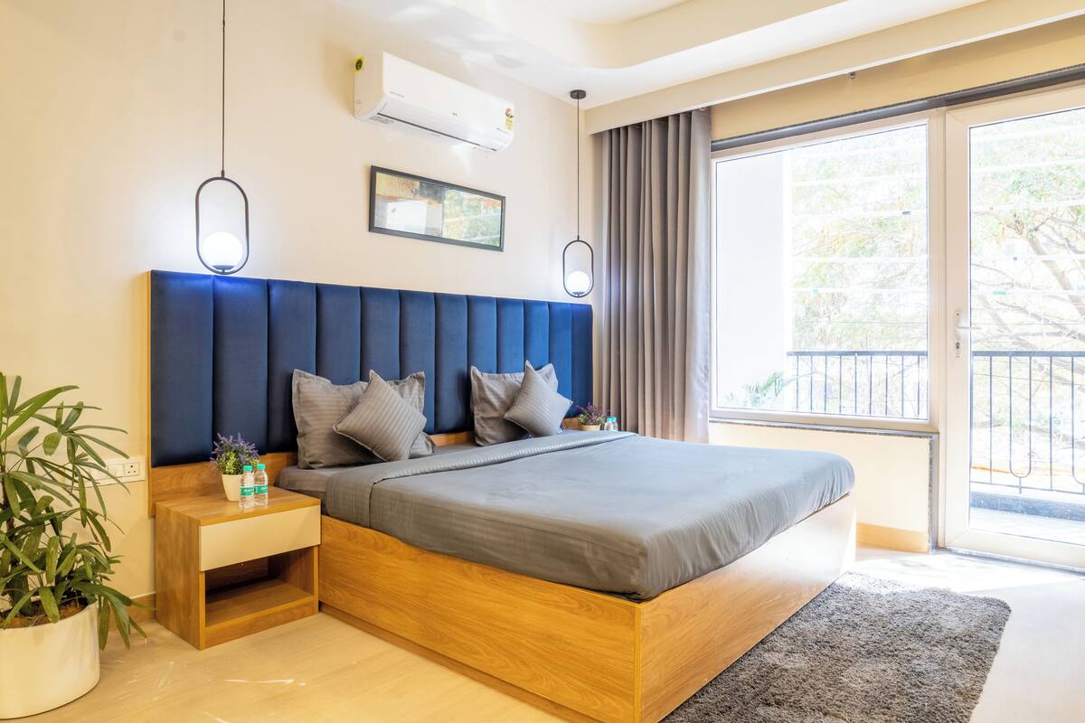 A modern bedroom with a large bed, wooden headboard, bedside tables, a potted plant, and a view of the outdoors.