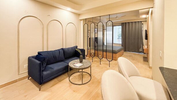 A modern living room with a blue sofa, a round coffee table, and a white armchair. A bed with a gray mattress and a view of the outdoors is visible in the background. - Housr 1 BHK Apartment Sector 42 Gurgaon (Gurugram)