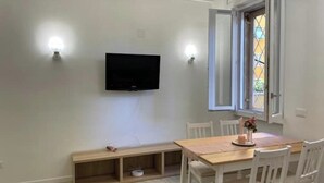 Uma sala de jantar com uma mesa de madeira e cadeiras brancas, uma televisão na parede e uma janela com vista para o exterior.