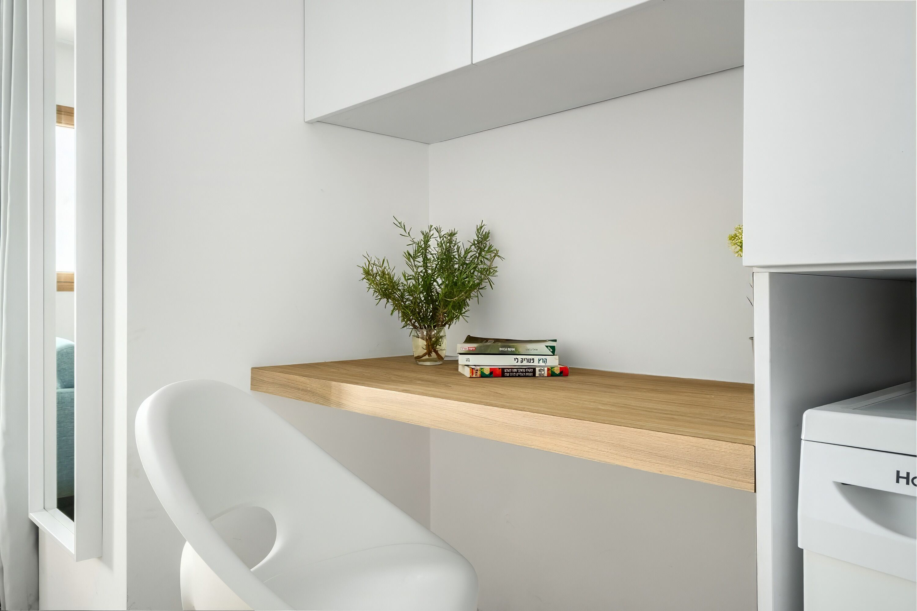 A modern, minimalist room with a white chair, a wooden shelf with books and a small plant, and a white cabinet.