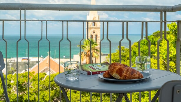 Varanda com mesa posta para refeição, vista para a costa com uma torre de igreja e vegetação.