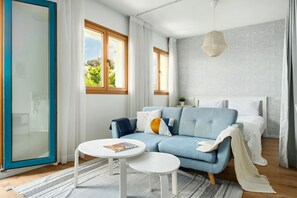 A modern bedroom with a blue sofa, a white bedside table, a bed with white bedding, and a large window with curtains.