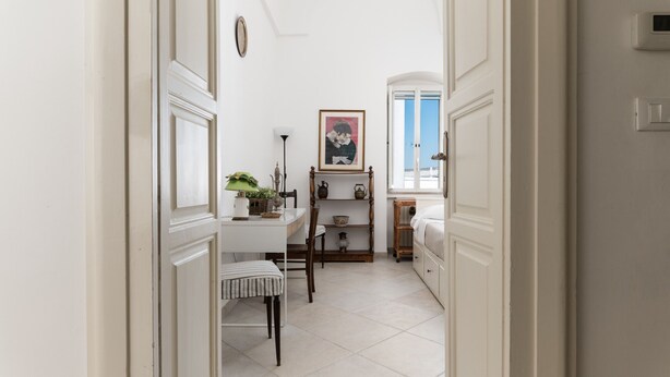 Un pasillo estrecho con una puerta blanca a la izquierda, una mesita con una lámpara y una planta, y una silla.