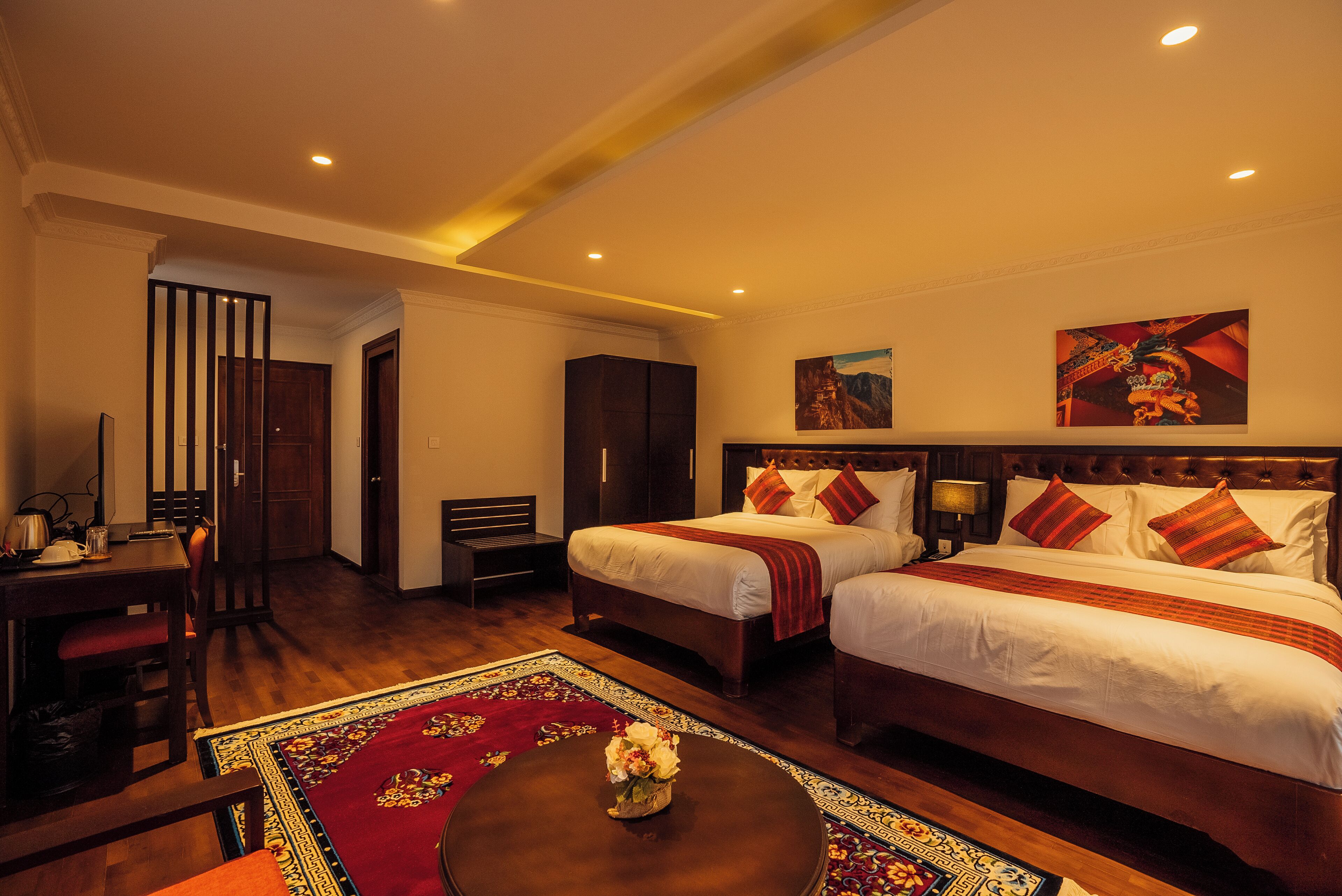 A hotel room with two beds, a desk, a chair, a small table with a vase, and a decorative rug.