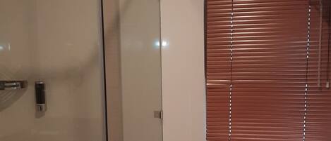 Un baño con ducha envuelta en vidrio, un inodoro y una ventana con persianas.