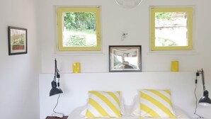 Un dormitorio con dos ventanas, una cama con almohadas a rayas amarillas y blancas y una lámpara de noche.