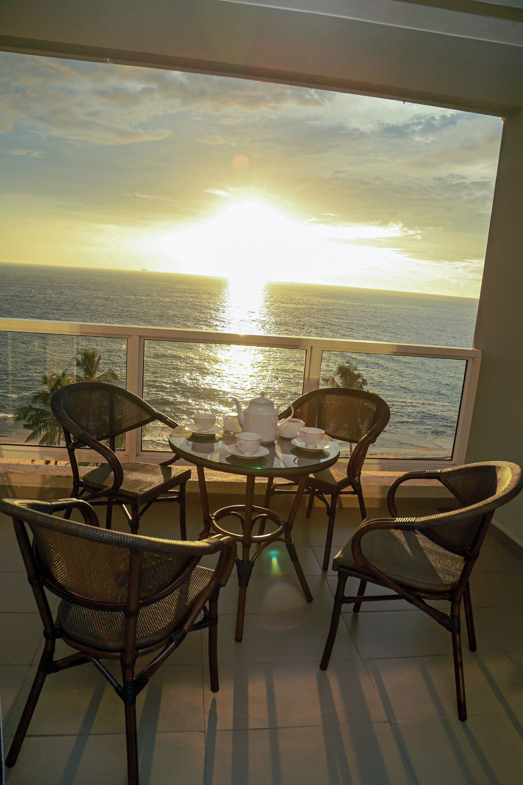 A balcony with wicker chairs and a table set for tea, overlooking a sunset over the ocean.