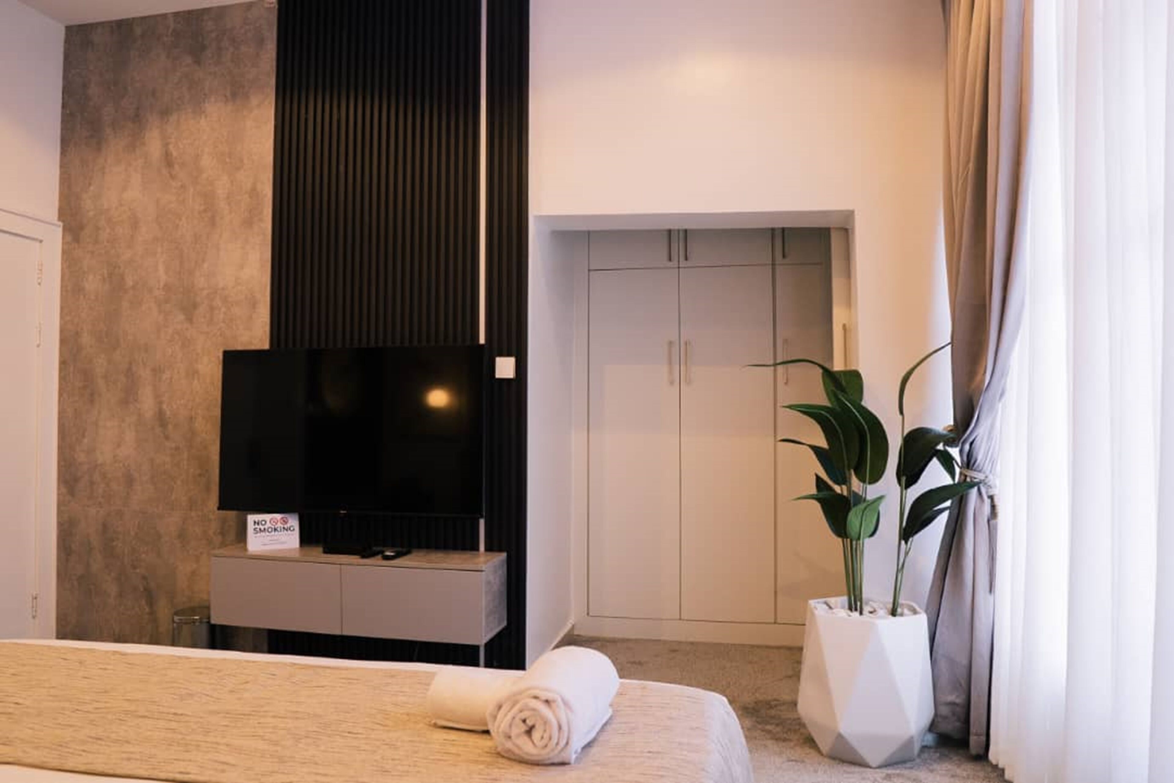 A modern hotel room with a large bed, a flat-screen TV, a wardrobe, and a potted plant.