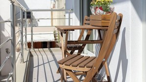 Une chaise et une table en bois sur un balcon doté d’une balustrade blanche, d’une fenêtre et offrant une vue sur des bâtiments en arrière-plan.