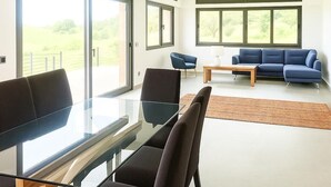 En moderne stue med et glasbord, mørke stole, en blå sofa og store vinduer med udsigt til det fri.