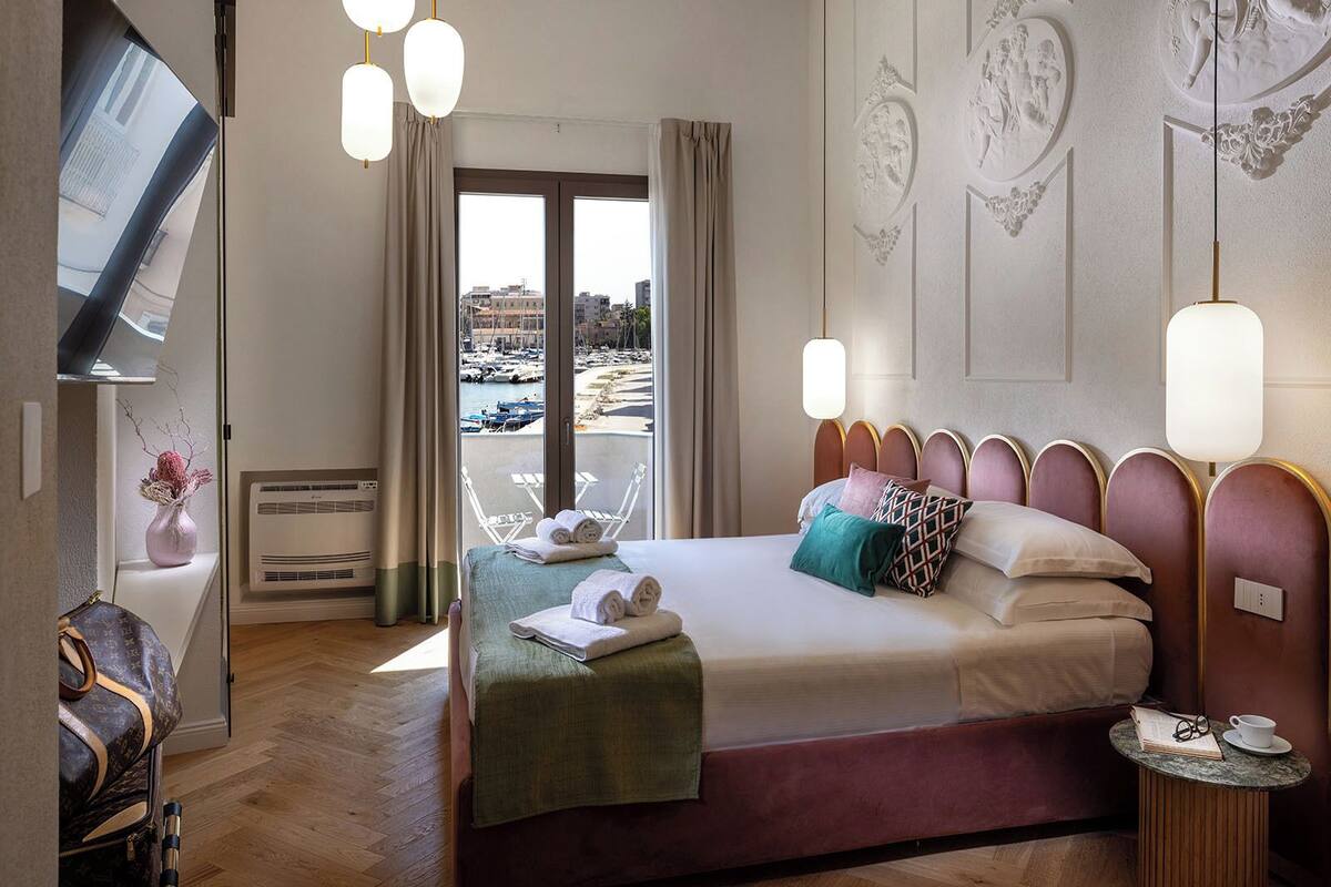 Une chambre d’hôtel avec un lit, une télévision, une chaise, une table avec un livre et des lunettes, et une vue sur la mer et les bateaux.