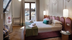 Une chambre d’hôtel avec un lit, une télévision, une chaise, une table avec un livre et des lunettes, et une vue sur la mer et les bateaux.
