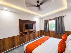 Een hotelkamer met een bed, een televisie bevestigd aan een houten paneel, een plafondventilator en een raam met gordijnen.