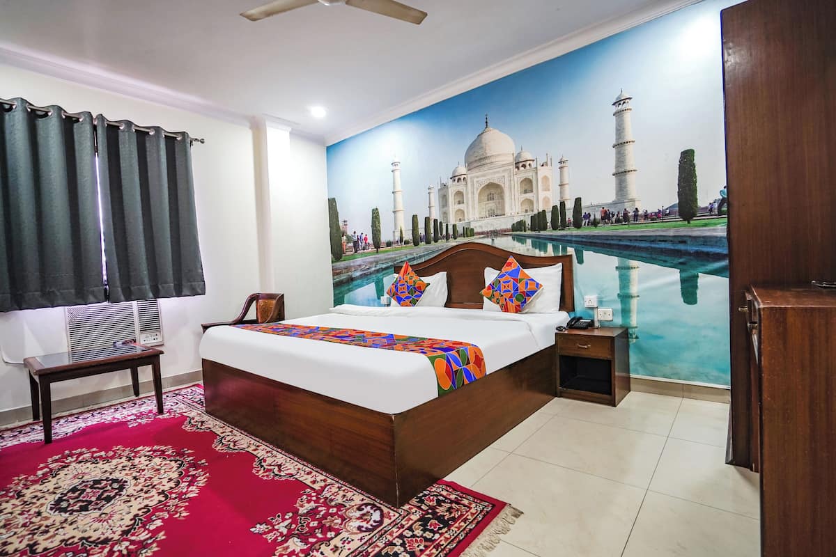 A bedroom with a large bed, a nightstand, a wooden wardrobe, a red patterned rug, and a wall mural of the Taj Mahal.