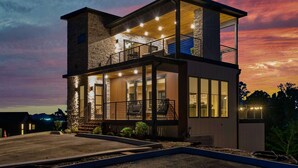 A modern two-story house with a balcony, large windows, and a well-lit exterior at dusk.