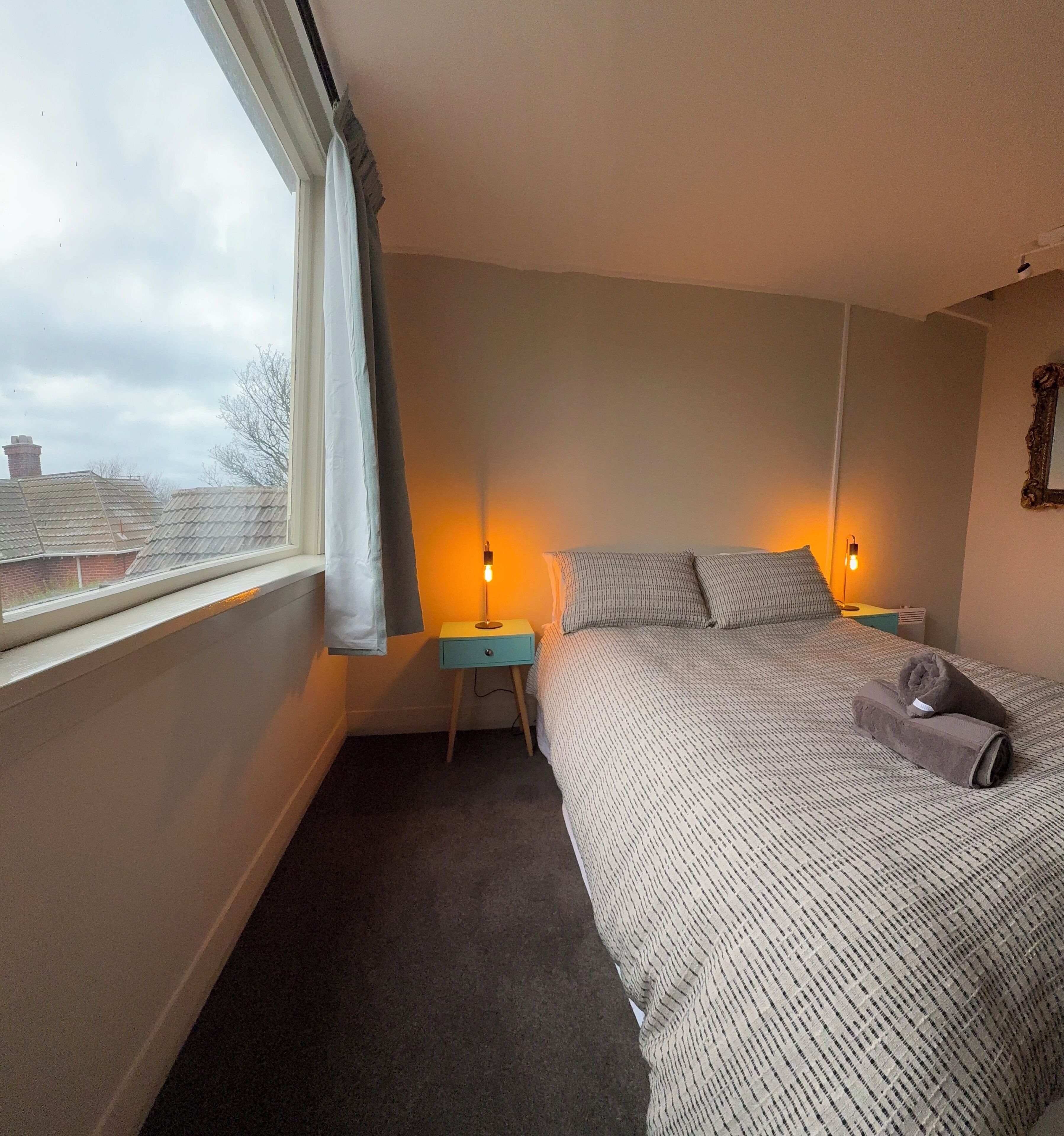 Un dormitorio con cama, dos mesitas de noche, una ventana con cortinas y vistas a edificios.
