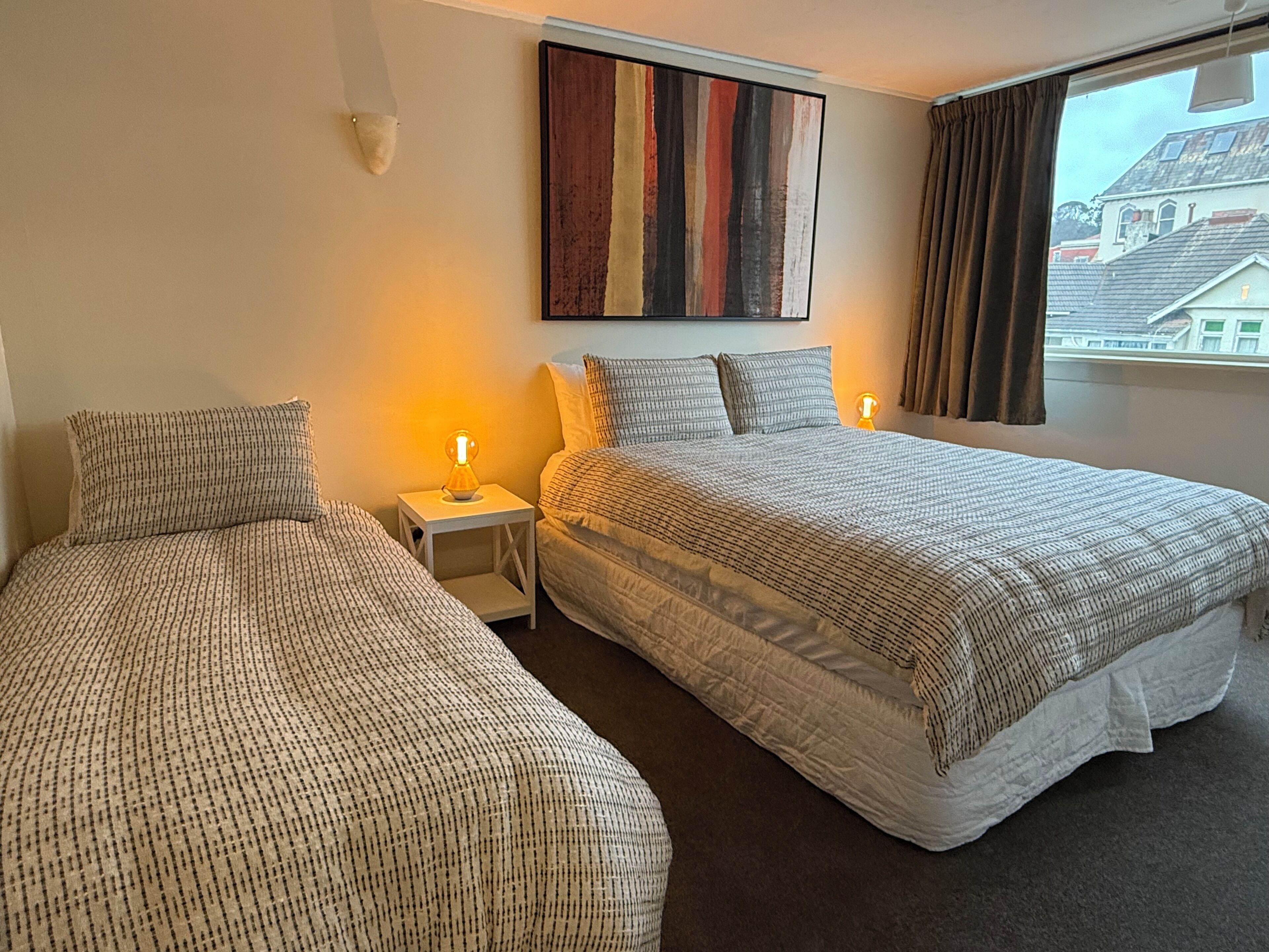 Habitación de hotel con dos camas, una mesita de noche, un cuadro en la pared y una ventana con cortinas.