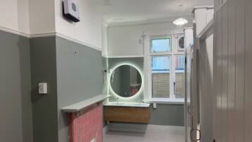 A bathroom with a white vanity, a round mirror with lighting, a window with blinds, and a door with a handle.