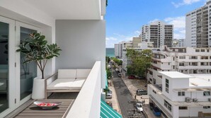A balcony with a white sofa and a potted plant, overlooking a street with buildings and a clear sky. - Sun Drift Views | Modern in the Heart of Condado (San Juan)