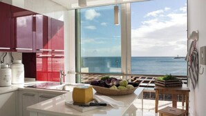 A modern kitchen with a large window overlooking the sea, a wooden stool, and a basket of fruits. - Boavista Apartment (Praia da Vitória)